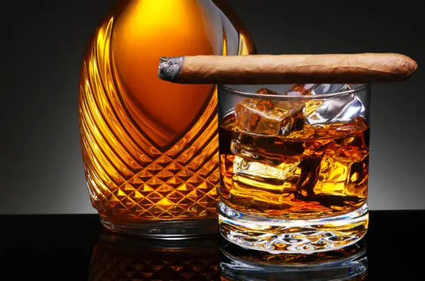 Closeup of an elegant decanter and a glass of scotch on the rocks with a lit cigar laying across the top of the glass. Horizontal on a light to dark gray background.
