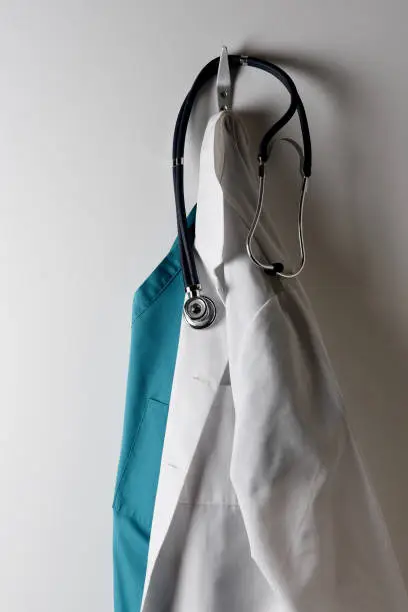 A Doctors White Lab Coat and Green Scrubs Hanging on a Hook with Stethoscope.