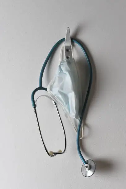 Closeup of a stethoscope and surgical mask hanging from a hook on a wall.
