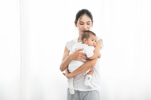 Pretty asian woman holding a newborn baby in her arms. Happy family. Laughing mother lifting her adorable infant baby son on white.