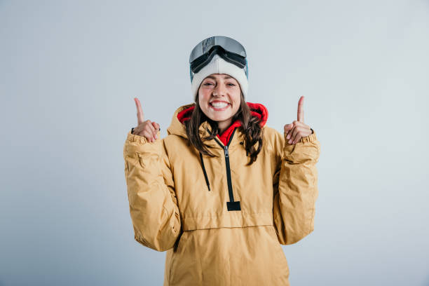 A cute girl standing in the studio, pointing with both fingers up to the sky, wearing all warm clothes, ready for the snow/skiing with a cap, and ski goggles on the head. Wearing a big smile radiating excitement!