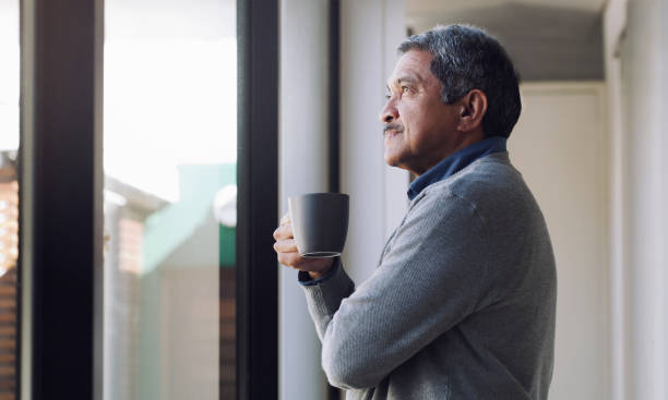 There's always something to look forward to Shot of a senior man drinking coffee and looking thoughtfully out of a window 65 69 years stock pictures, royalty-free photos & images