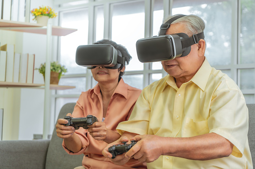 Senior Asian couple is playing Video game and wearing VR goggles with joypad for retirement technology lifestyle concept.