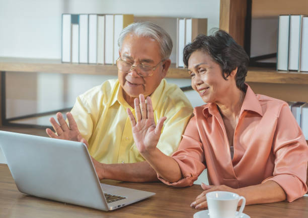 istock Senior Mature Asian couple is waving to computer while using video calling technology to talk to family from home. 1283631833