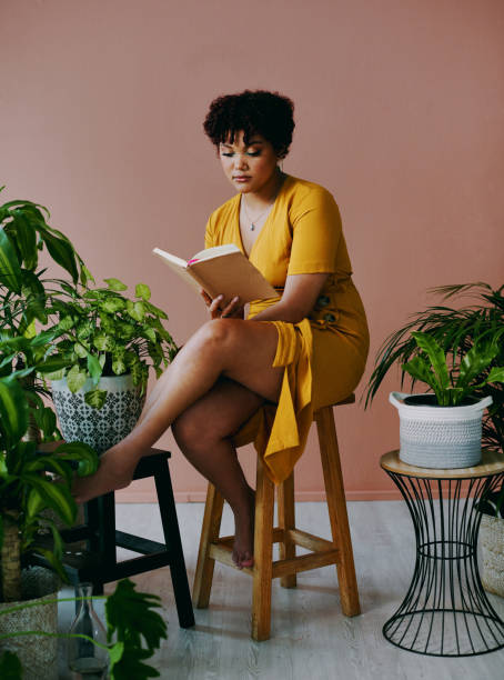 Shot of a young woman reading a book while sitting with plants around her at home