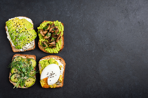 Set of avocado toasts with different toppings. Vegetarian avocado toast with egg, fried mushrooms, cucumber, seeds and micro greens on a black concrete background. Top view