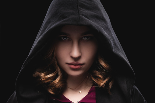 Close-up portrait of a secretive young girl in a deep dark hood on a black background. The concept of secrecy of secrets and people hiding from the government. Hackers and thieves. Low key