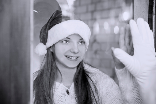 Teenage Girl holding hand on window glass touching Santa Claus palm hand on the other side. Black and white portrait of cute girl touch Santa through window