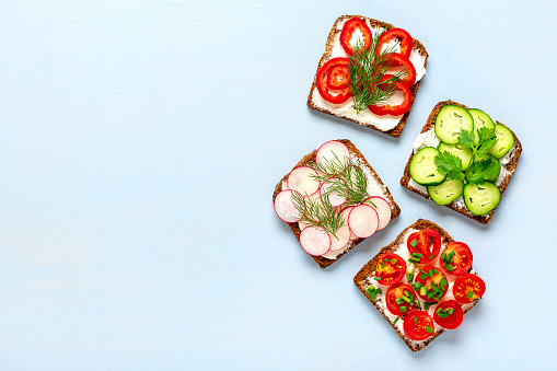 Variety of sandwiches for breakfast - slice of whole grain dark bread, pepper, cream cheese, cucumbers, radishes, cherry tomatoes, garnished with dill, green onions on blue table Top view Flat lay.