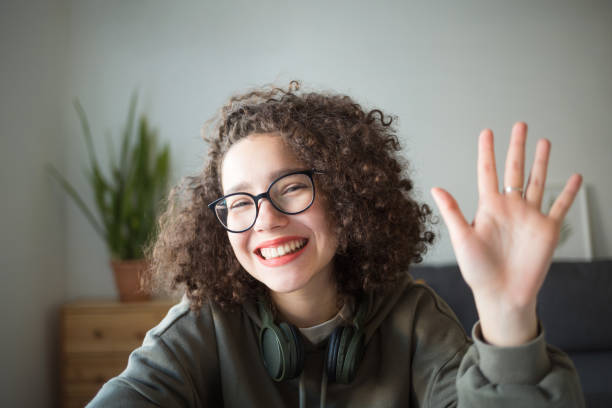 girl looks at camera, waves her hand and smiles. student talks over Internet.