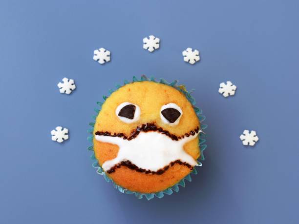 Homemade muffin with protective face mask on blue background, small sugar snowflakes around
