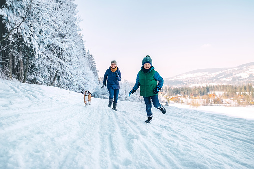 Mother and son having a fun during dog walk. They running with their beagle dog in snowy forest. Mother and son relatives and femily values concept image.
