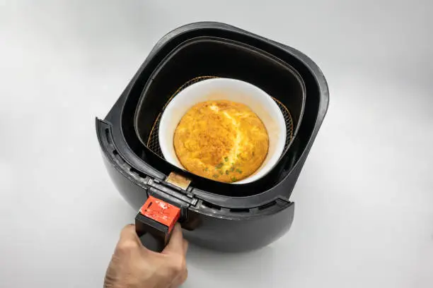 Photo of Omelet in white ceramic bowl within black air-fryer isolated on white background.