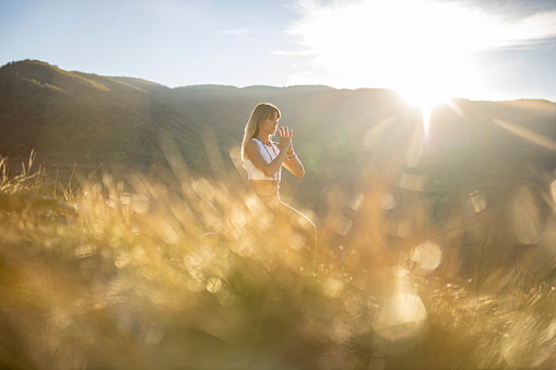 Shot of one woman in nature practicing yga. Lens flare, soft light, grass and mountains on the back are making serene scene.