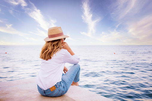 Full length shot of woman wearing sun hat and white shirt with blue jeans while sitting on seaside and daydreaming.