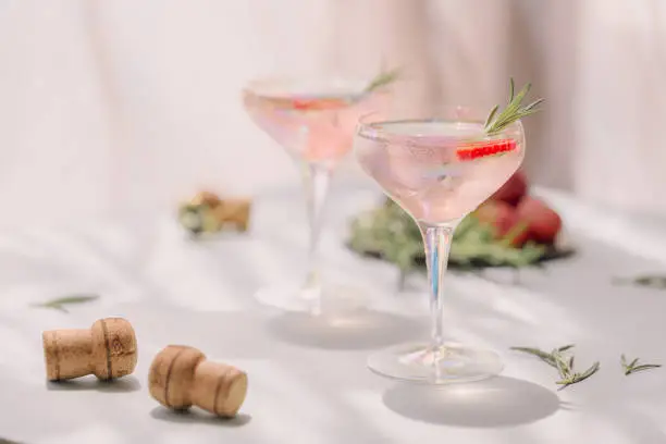 Photo of Summer drink with white sparkling wine. Homemade refreshing fruit cocktail or punch with champagne, strawberries, ice cubes and rosemary on beige sandy background.