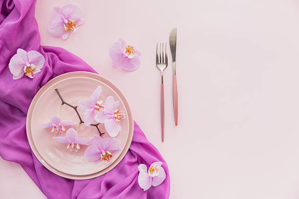 istock Flower and table settings overhead composition on light pink background. Pink ceramic plates, cutlery and perple orchid flowers. Holiday modern table decoration. Romantic date. 1283685319