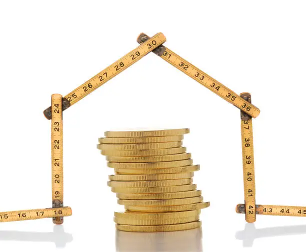 High Cost of Housing Concept: A carpenters ruler in the shape of a house with a stack of gold coins.