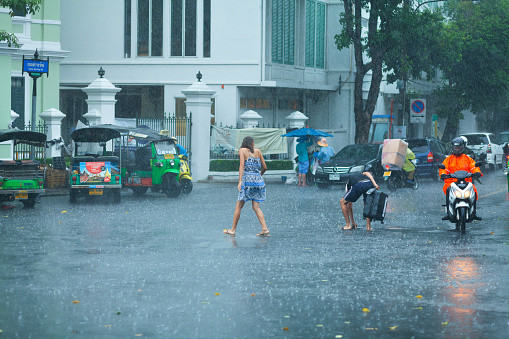 Caucasian tourist couple captured in hard rain in Bangkok. Couple is crossing street and man has lost a sandal in water. At right side a motorcycle is driving. In left background tuktuks are parked