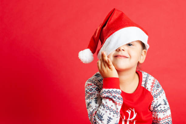 крупным планом улыбающийся ребенок в рождественской пижаме, скрывая один глаз под шапкой санта-клауса, копировать пространство на красном  - скрывая стоковые фото и изображения
