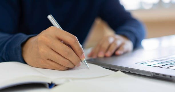 istock close up man hand using pen to writing homework or practice exam before test while opening laptop to learning from webinar for e-learning new normal lifestyle concept 1283690891