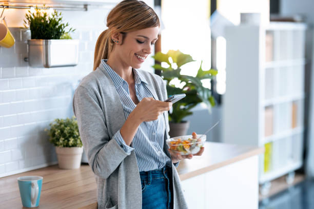 belle jeune femme utilisant son téléphone portable tout en prenant le petit déjeuner sain dans la cuisine à la maison. - oatmeal breakfast healthy eating food photos et images de collection
