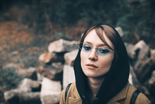 pretty young girl in olive long coat, hooded sweatshirt, backpack and glasses. Portrait of woman with hood, Stones on a background