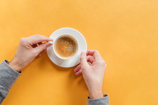 Man's hands holding a cup of coffee on yellow table, top view