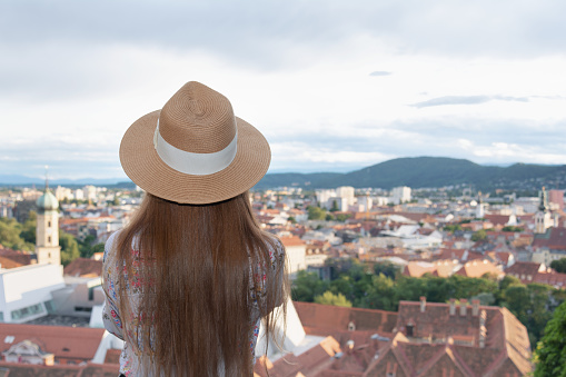 A girl with long hair in a hat stands on the observation deck and looks at the city of Graz. Travel concept, architecture, people.