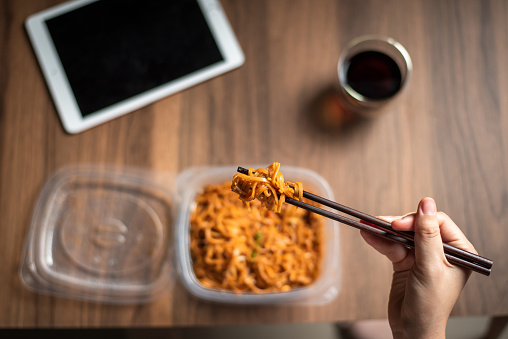 Woman having takeaway or delivery Pad Thai fried noodles with chicken meal from a plastic package with juice at home first person view