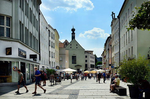 Passau, Germany - July 17, 2018: unknown people in Ludwigstrasse in the old town with different shops and Marist monastery with a distinctive onion dome, the city lies at the confluence of the Danube, Ilz and Inn rivers and is therefore also called the three rivers city