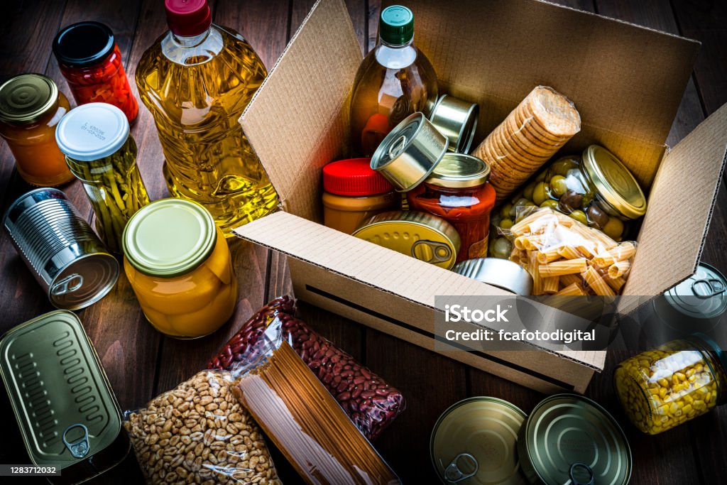 Cardboard box filled with non-perishable foods on wooden table. High angle view. High angle view of a cardboard box filled with multicolored non-perishable canned goods, conserves, sauces and oils shot on wooden table. The composition includes cooking oil bottle, pasta, crackers, preserves and tins. High resolution 42Mp studio digital capture taken with SONY A7rII and Zeiss Batis 40mm F2.0 CF lens Food Stock Photo