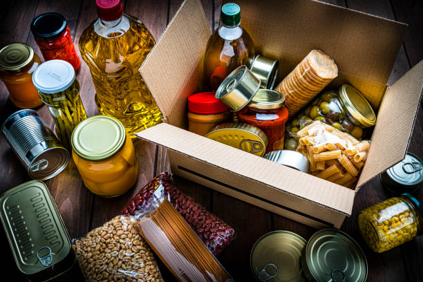 Cardboard box filled with non-perishable foods on wooden table. High angle view. High angle view of a cardboard box filled with multicolored non-perishable canned goods, conserves, sauces and oils shot on wooden table. The composition includes cooking oil bottle, pasta, crackers, preserves and tins. High resolution 42Mp studio digital capture taken with SONY A7rII and Zeiss Batis 40mm F2.0 CF lens cardboard box photos stock pictures, royalty-free photos & images