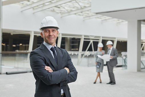 Waist up portrait of smiling mature businessman wearing hardhat and looking at camera while standing with arms crossed at construction site, copy space