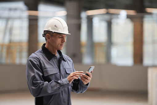 Side view portrait of mature worker using smartphone while standing at construction site or in industrial workshop, copy space