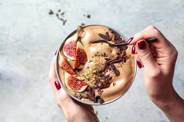 Photo of Woman holding a bowl of chocolate smoothie with figs and hemp seeds, gray background. Healthy vegan food concept.