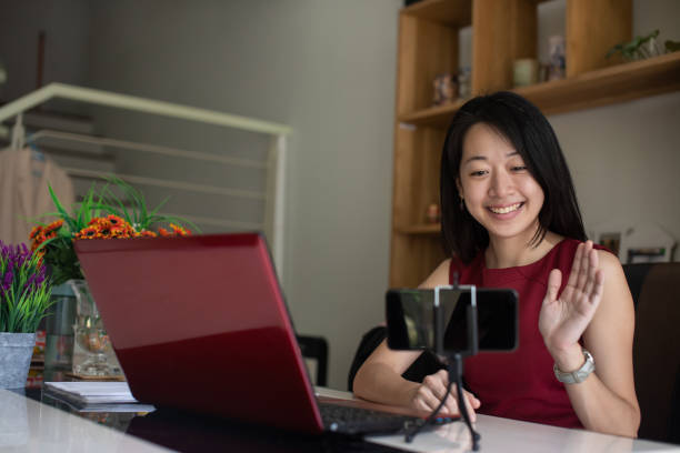 若いアジアの起業家は、mco(移動制御命令)期間中に自宅で自分のワークステーションでノートパソコンを扱っている間、スマートフォンで顧客とビデオチャットを持っています。 - m&co ストックフォトと画像