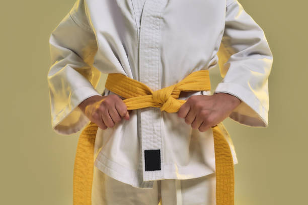tiro cropped de menina karatê em quimono branco ajustando cinto amarelo enquanto se prepara para se exercitar, fazer artes marciais, ficar isolado sobre fundo amarelo - karate child judo belt - fotografias e filmes do acervo