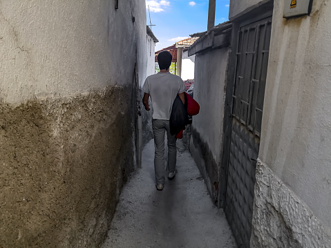 Turkey, Ankara - October 23, 2019: A tall man walks along a narrow aisle between two shabby walls outdoors, view from the back. Male silhouette of a person in a white t-shirt and light trousers on a narrow urban path