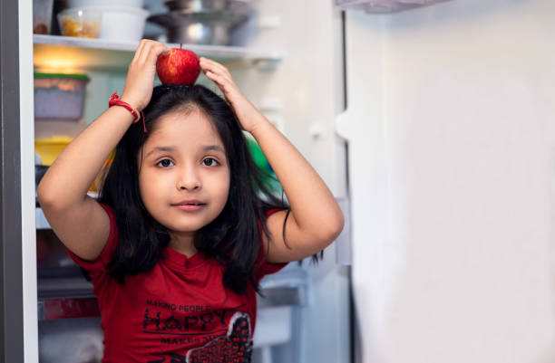 милая маленькая девочка, держащая яблоко и стоящая возле холодильника. - refrigerator healthy eating mango fruit стоковые фото и изображения