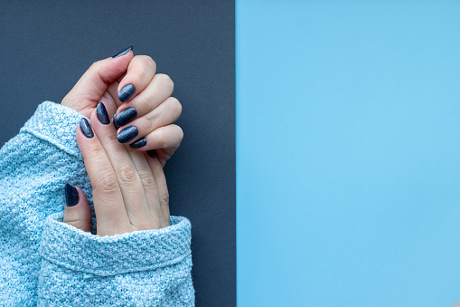 Female hands in knitted sweater fabric with beautiful manicure - dark glittered nails on gray and blue background with copy space