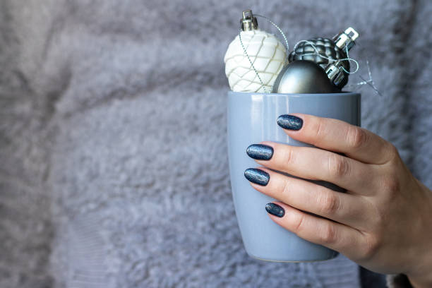 Female hand in gray fluffy knitted sweater with beautiful manicure - dark gray blue glittered nails with cup with Christmas decorations with copy space Female hand in gray fluffy knitted sweater with beautiful manicure - dark gray blue glittered nails with cup with Christmas decorations with copy space christmas nails stock pictures, royalty-free photos & images