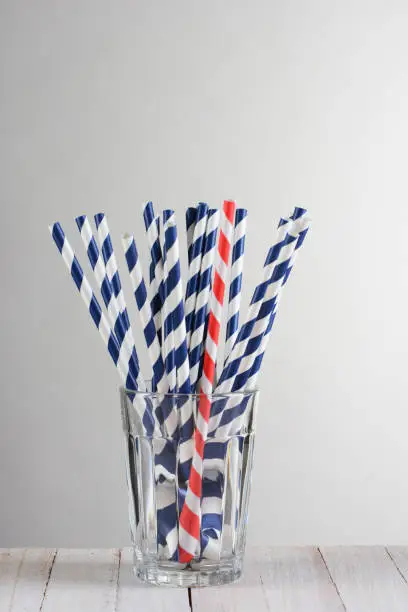 One red striped drinking straw mixed in with a bunch of blue striped straws. The straws are fanned out in a drinking glass on a wood table against a light to dark gray background.