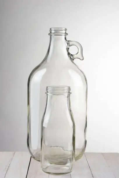 Two empty glass bottles, a small bottle in front of a larger container. Vertical format against a light to dark gray background. The bottles are set on a rustic wood table.