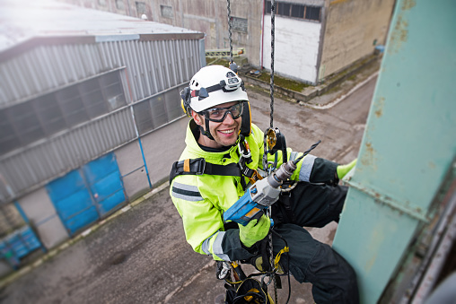 Manual rope access technician - worker repairing tower - antenna in sunshine, holding drill in hand, preparing for work and looking to camera and smiling