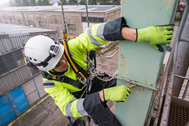 Manual rope access technician - worker repairing tower - antenna in sunshine, holding screw in hand and tightens the screws. Industrial buildings behind stock photo