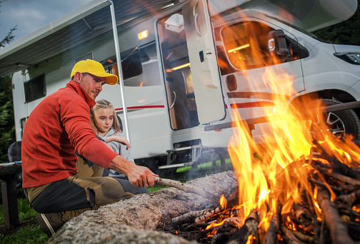 Recreational Vehicle RV Camper Camping and Family Time. Caucasian Father and His Daughter Hanging Next to Campfire on Their RV Park Pitch. Class C Motorhome in Background.