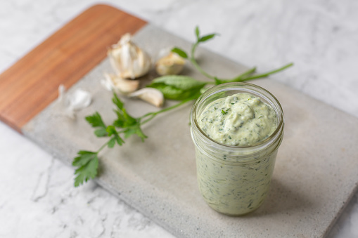 A small jar of homemade green goddess dressing on a cement cutting board on a white marble countertop.  A sprig of parsley and several cloves of garlic are in the background.