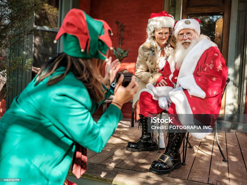 Santa and Mrs.Clause pose for photos for Elf Santa Claus and Mrs.Clause pose for photos onutdoors on th porch while Elf is directing in foreground with the camera. Santa Claus Stock Photo