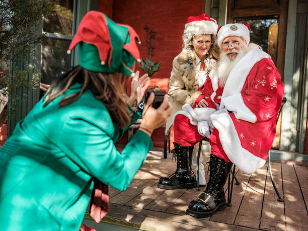 Santa and Mrs.Clause pose for photos for Elf Santa Claus and Mrs.Clause pose for photos onutdoors on th porch while Elf is directing in foreground with the camera. elf photos stock pictures, royalty-free photos & images
